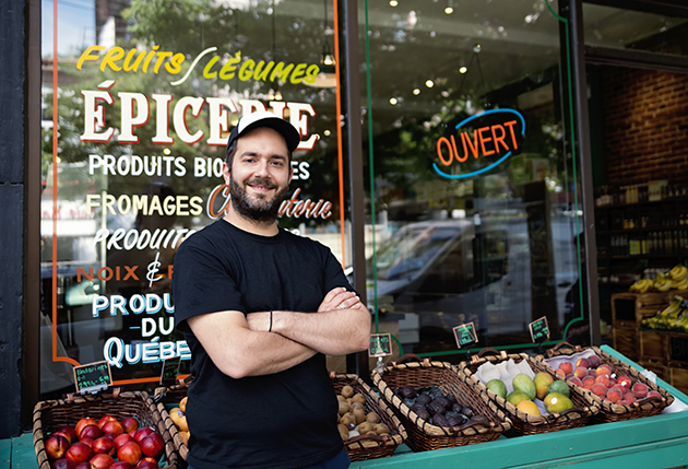 Un homme en tee-shirt noir, les bras croisés, pose devant l'étalage de fruits et légumes d'une épicerie québécoise