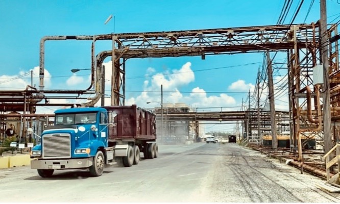 A large garbage truck with a dumpster on the back driving through an industrial facility