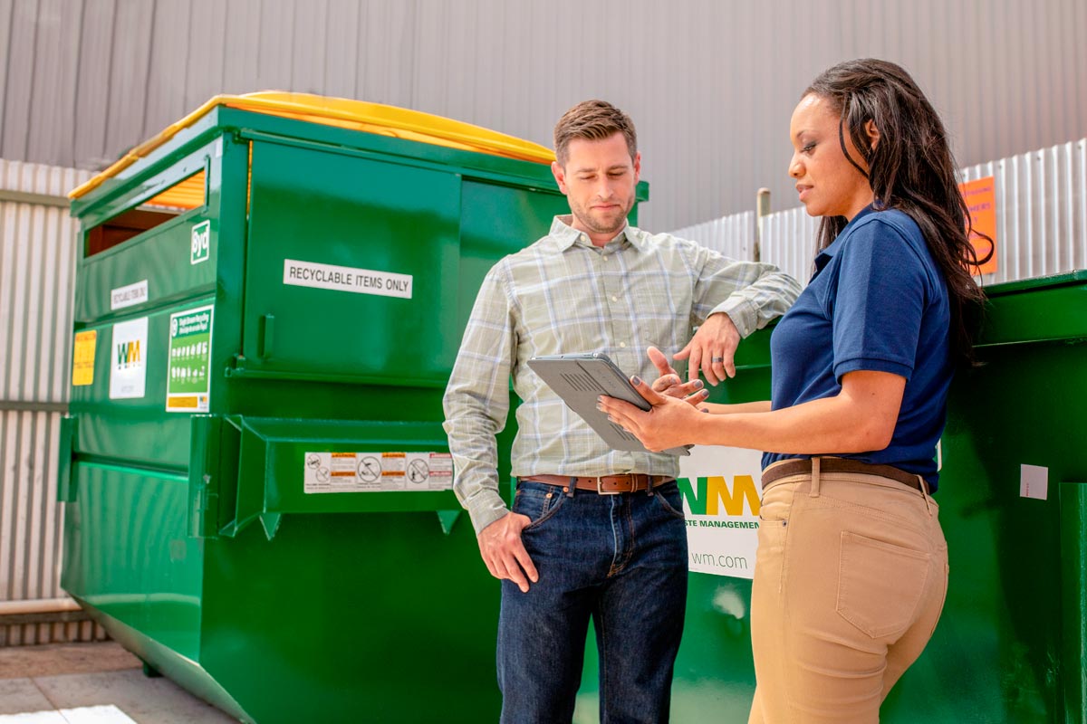 A man and woman look at a tablet while standing next to a WM dumpster.