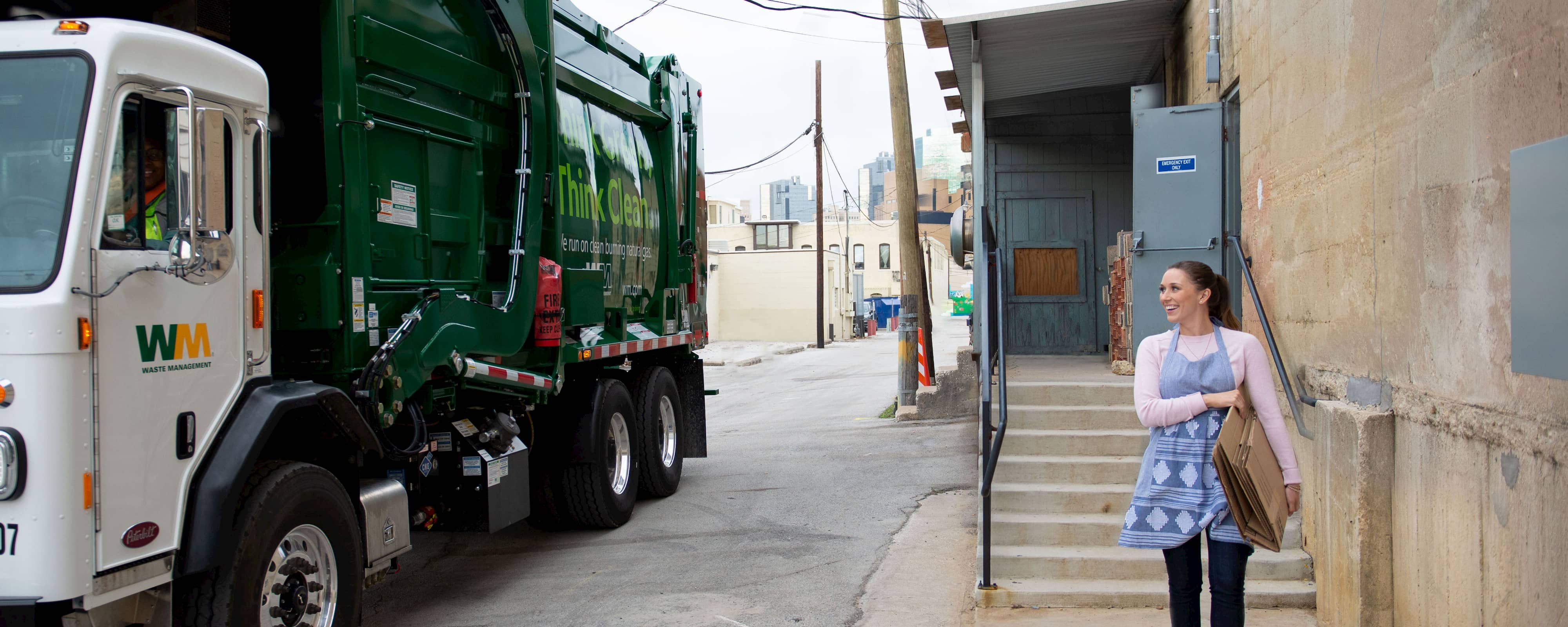 Woman in an apron carrying cardboard boxes out to a Waste Management dumpster truck.