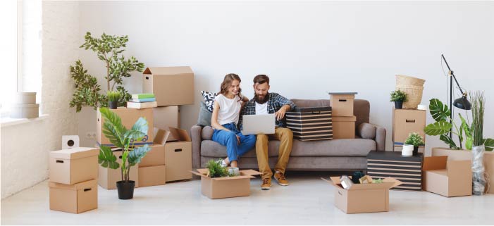 A couple sits on a couch surrounded by boxes while looking at a laptop computer.