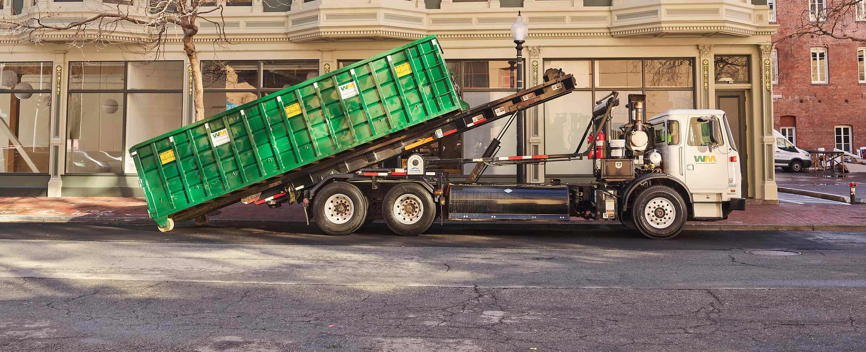 Small semi truck hauling large green rental dumpster down city street in front of local businesses