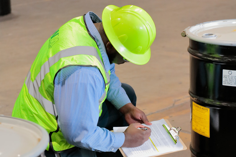 Technician kneeling and taking notes from a barrel in a warehouse while wearing a safety vest