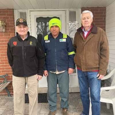 Younger WM male employee standing on the porch of a brick suburban home with two older men