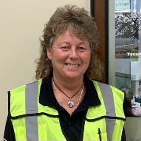 Sanitation worker with tall curly hair standing in office building with black collared shirt and green safety vest