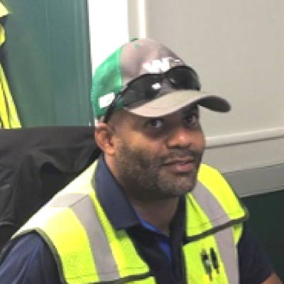 Smiling sanitation worker with beard wearing mesh baseball cap, sunglasses, collared uniform shirt, and green safety vest