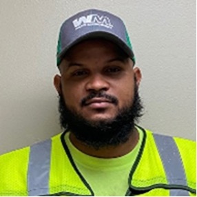Bearded WM employee standing in front of white wall wearing mesh baseball cap, green t-shirt, and a reflective safety vest