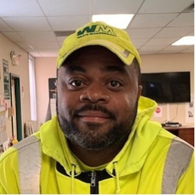 Older male waste management worker with greying beard sitting inside office wearing brightly colored safety clothing