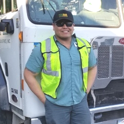 Waste collection worker in sunglasses, hat, and safety vest leaning against front of WM garbage truck