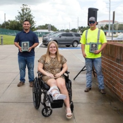 Two sanitation workers- one in a safety vest and the other in a blue polo, both holding plaques and standing next to a wheelchair user