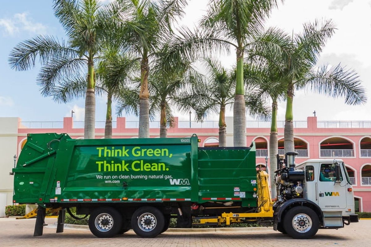 A WM garbage pickup truck parked in front of some palm trees in Durant, Florida and a pink building in the background.