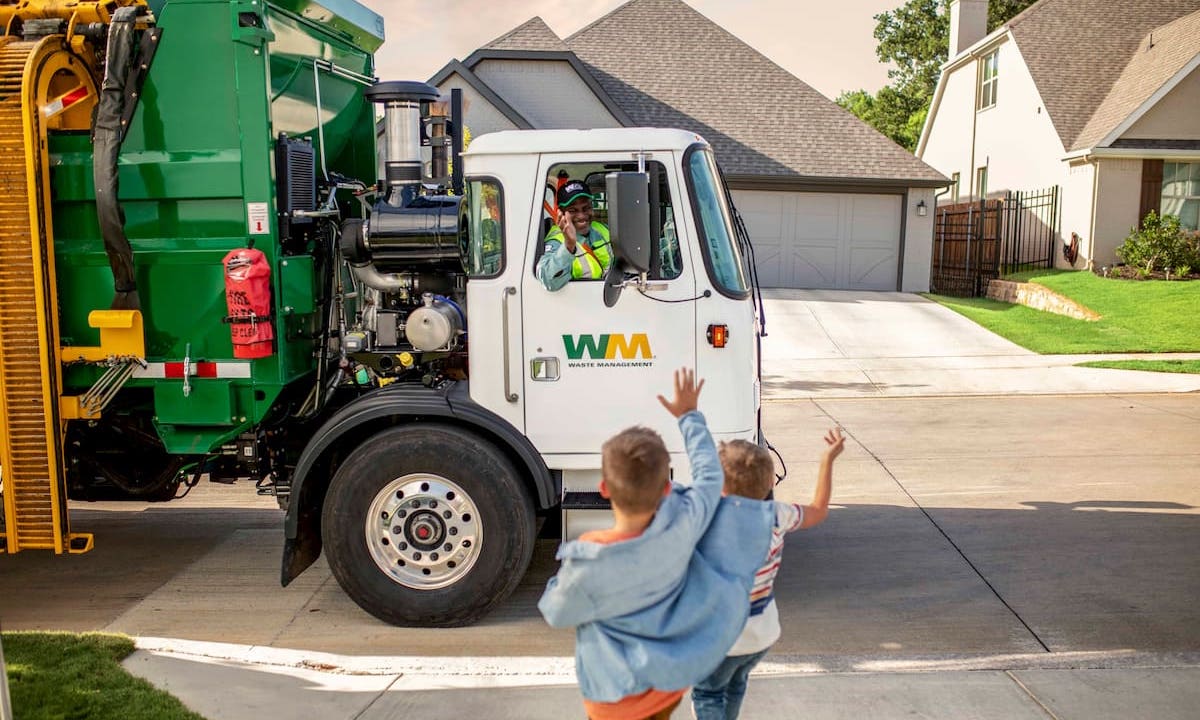 WM garbage truck passing through suburban neighborhood while driver smiles and waves to two nearby children